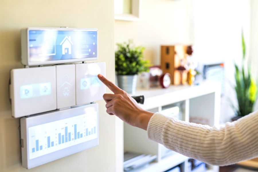 Une main pointant vers un panneau mural modulaire avec écrans tactiles affichant des données de maison connectée, au cœur d'un système de gadgets connectés domotiques.