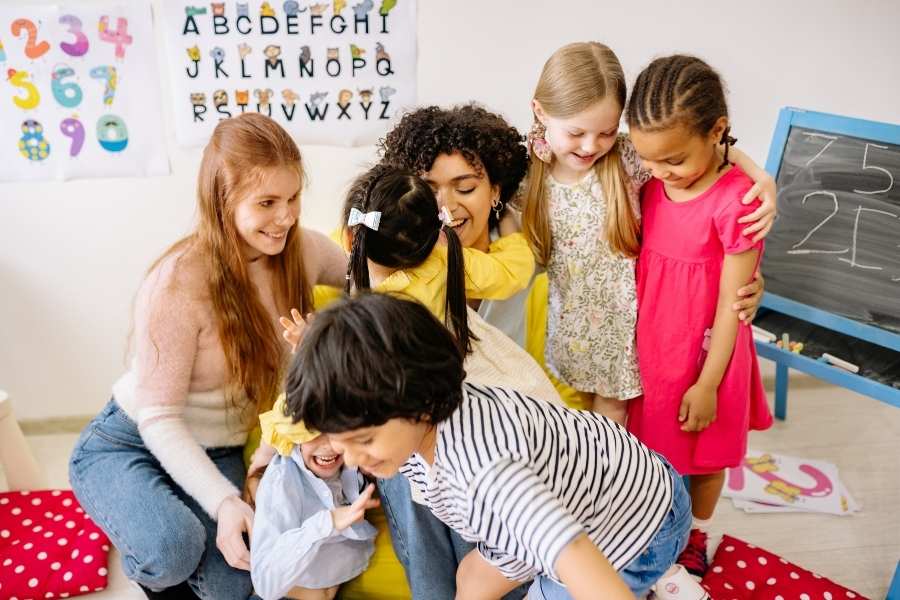 Un groupe d'enfants et deux adultes s'enlacent joyeusement dans une salle de classe, illustrant l'importance des interactions humaines réelles face aux dangers cachés des réseaux.